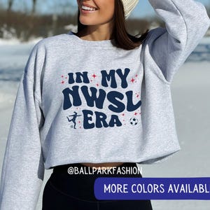Op de afbeelding: Een lichtgrijs sweatshirt met de tekst "IN MY NWSL ERA" in marineblauw en rood. Het ontwerp bevat een voetballer en een voetbal. De sweatshirt heeft een ronde hals en lange mouwen.