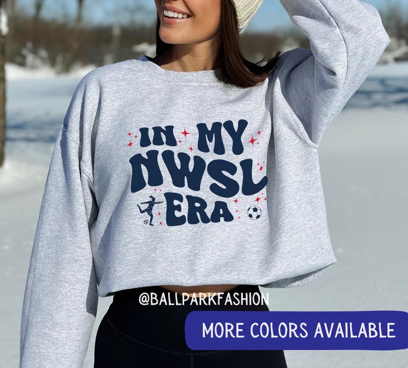 Op de afbeelding: Een lichtgrijs sweatshirt met de tekst "IN MY NWSL ERA" in marineblauw en rood. Het ontwerp bevat een voetballer en een voetbal. De sweatshirt heeft een ronde hals en lange mouwen.