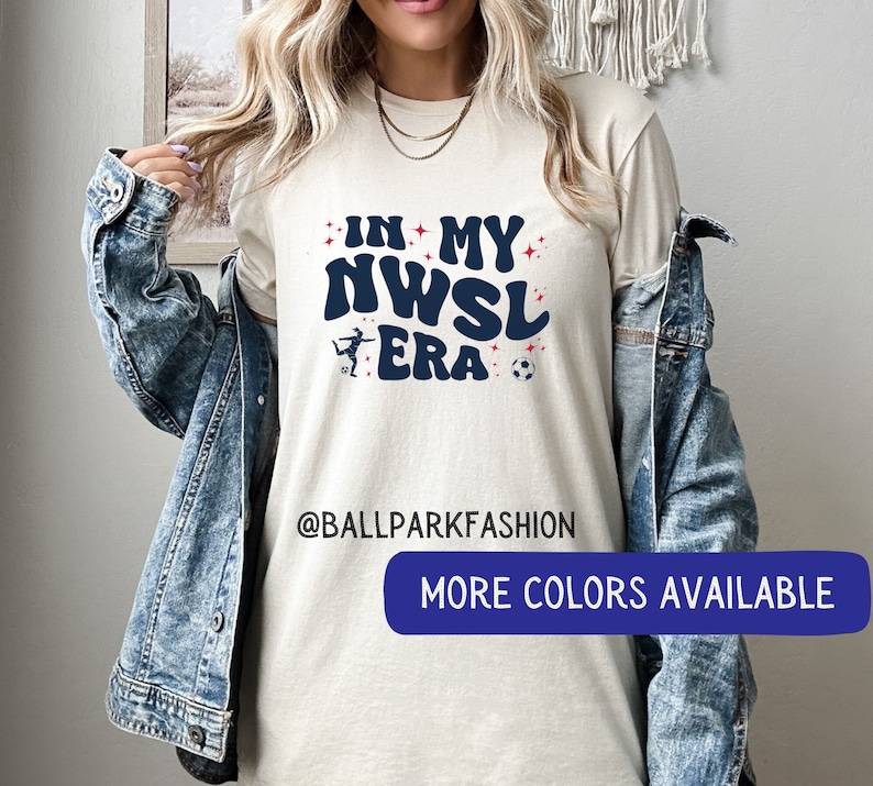 Pu&ograve; includere: Una maglietta bianca con il testo "In My NWSL Era" in lettere blu e rosse. Una silhouette di un calciatore &egrave; presente nel design. Il testo "@ballparkfashion" &egrave; stampato sotto il design. Il testo "Altri colori disponibili" &egrave; stampato in blu su un rettangolo bianco.