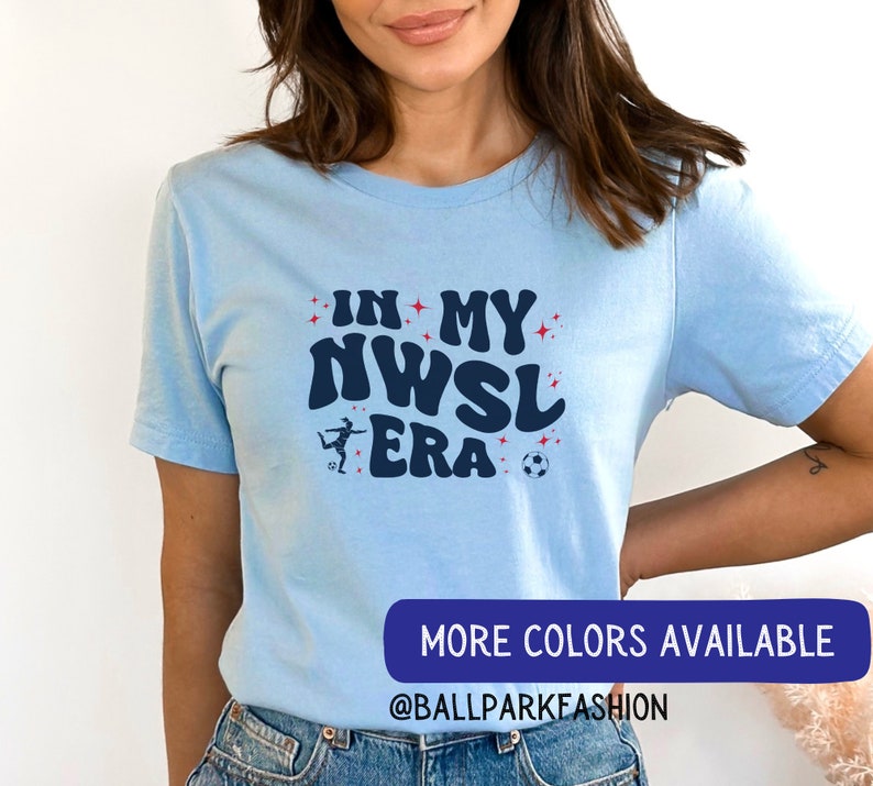 Pu&ograve; includere: Una maglietta blu chiaro con il testo "In My NWSL Era" in un font retr&ograve;. &Egrave; mostrata una silhouette di un calciatore che calcia un pallone da calcio. Il testo "Altri colori disponibili" e "@ballparkfashion" sono mostrati nella parte inferiore dell'immagine.