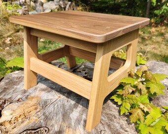 May include: A small wooden stool featuring a rectangular top with a dark brown wood grain finish and a lighter tan wood base. The stool has four sturdy legs connected by horizontal support beams, creating an open frame design with rounded edges.