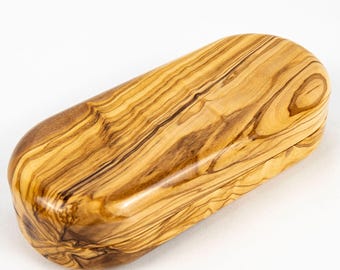 May include: An oblong, rounded wooden box crafted from olive wood, featuring a smooth, polished surface that highlights the natural, swirling grain patterns in shades of light tan, golden yellow, and deep brown throughout the entire exterior.