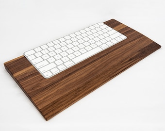May include: A white wireless keyboard sits on a wooden stand. The stand is made of dark brown walnut wood and has a slight incline. The keyboard has a white background with black keys and a silver frame.
