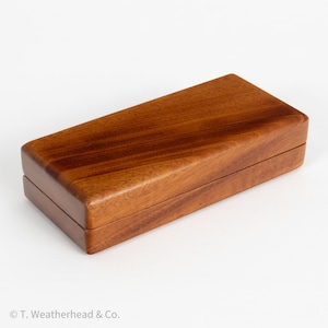 May include: A rectangular wooden box with a hinged lid. The box is made of a reddish-brown wood with visible grain patterns. The box is closed and sits on a white surface. The box is likely used for storage or display.