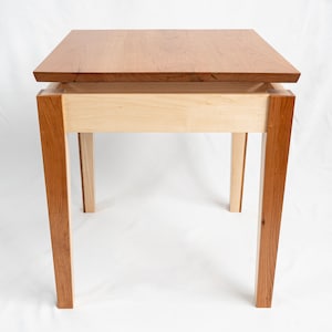 May include: A wooden end table with a cherry wood top and maple wood legs. The top is slightly raised above the legs, creating a unique design.