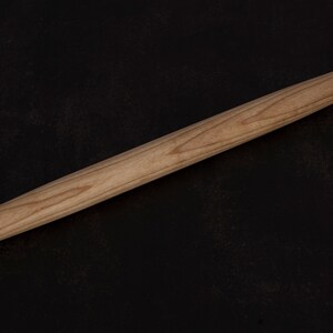 Puede incluir: Un rodillo de madera, de aproximadamente 45 cm de largo, con un acabado liso de color marr&oacute;n claro. El rodillo est&aacute; sobre una superficie negra.