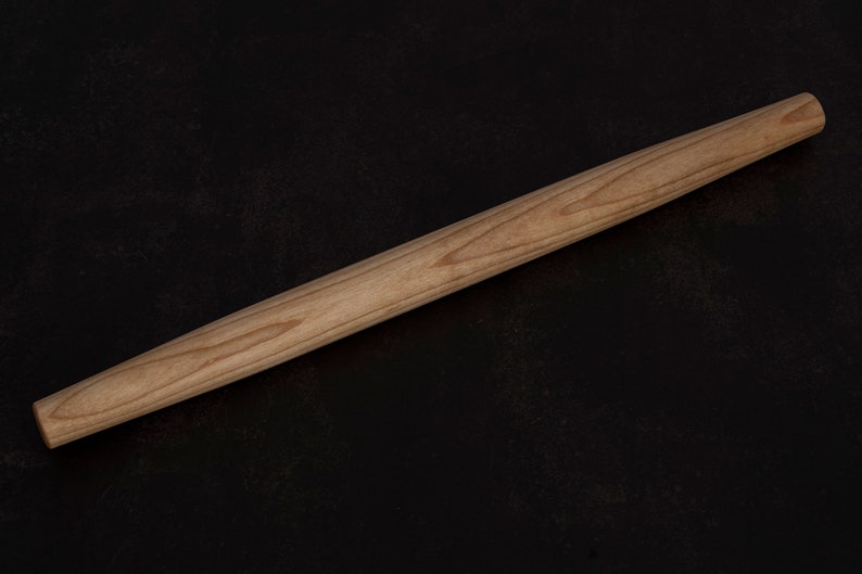 Puede incluir: Un rodillo de madera, de aproximadamente 45 cm de largo, con un acabado liso de color marr&oacute;n claro. El rodillo est&aacute; sobre una superficie negra.