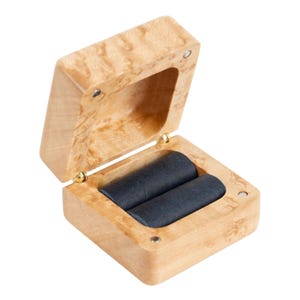 May include: Open wooden ring box with a light brown finish and a hinged lid. The interior features two black velvet ring slots. The box is made of wood with a natural grain pattern and gold-coloured hinges.