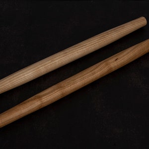 Puede incluir: Dos rodillos de amasar de madera sobre una superficie negra. Los rodillos de amasar son de madera marr&oacute;n claro y tienen un acabado liso.
