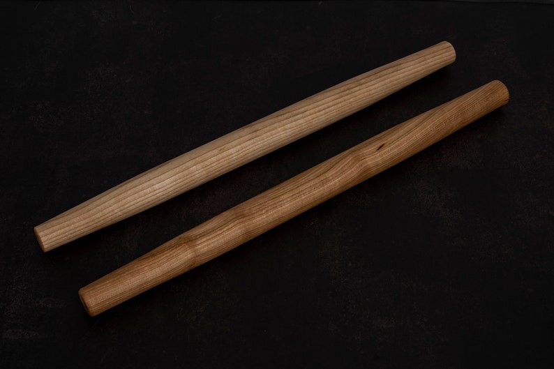 Puede incluir: Dos rodillos de amasar de madera sobre una superficie negra. Los rodillos de amasar son de madera marr&oacute;n claro y tienen un acabado liso.