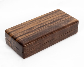 May include: A rectangular wooden box with a smooth, polished finish. The exterior displays a prominent dark brown wood grain pattern with lighter brown streaks running horizontally across the lid and the sides of the closed container.