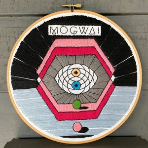 May include: Embroidered hoop art featuring a geometric design with a stylized eye in the center. The design is in shades of pink, red, gray, black, and white. The text "MOGWAI" is embroidered at the top of the hoop.
