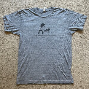 May include: A heather gray t-shirt with a black graphic of a person holding a record. The text on the shirt reads "I collect records because pussy hates me." The shirt is made of a soft material.