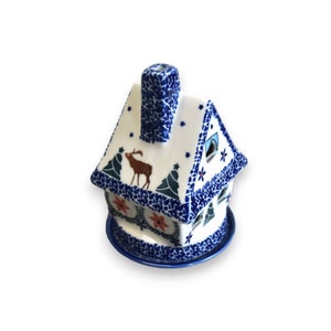 Può includere: Un portacandele in ceramica blu e bianca a forma di casa con un design di cervo e albero di pino. Il portacandele ha un camino e una base con un motivo floreale.