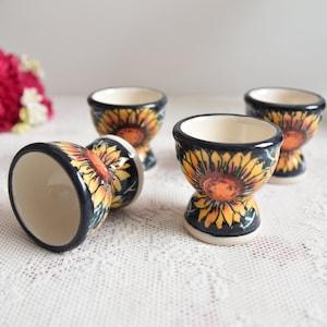 Può includere: Quattro porta uova in ceramica con un design a girasole. Le porta uova sono bianche con un bordo blu e presentano girasoli gialli e arancioni su uno sfondo blu scuro.