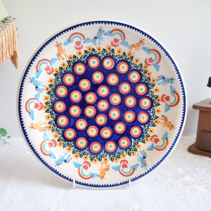 May include: A large, round ceramic plate with a blue and white peacock design. The plate features a central circle of blue and white concentric circles surrounded by a ring of smaller circles. The outer edge of the plate is decorated with a border of yellow flowers and rainbows.