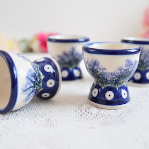 May include: Four blue and white ceramic egg cups with a floral design. The egg cups are on a white lace tablecloth.