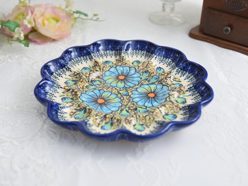 May include: A ceramic, scalloped-edge plate with a blue floral design. The plate features a dark blue rim and a cream-colored interior with blue and orange flowers. The plate is likely used for serving food.