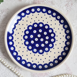 Pode incluir: Um prato de cerâmica branco com um padrão de bolinhas azuis e brancas. O centro do prato apresenta um design de círculo maior com anéis azuis e pontos brancos.