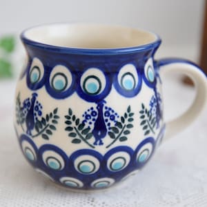 Può includere: Una tazza in ceramica bianca con un design di pavone blu e verde. La tazza ha un corpo arrotondato e un manico.