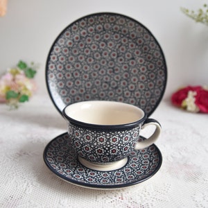 May include: A ceramic tea set featuring a cup, saucer, and plate. The set has a dark blue background with a repeating pattern of small red and white floral designs. The cup has a white interior and a curved handle.