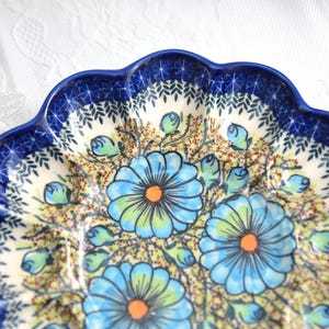 May include: A ceramic plate with a scalloped edge and a floral design. The plate features a dark blue rim with a pattern of leaves and a cream-colored center with blue flowers and green leaves. The flowers have orange centers.