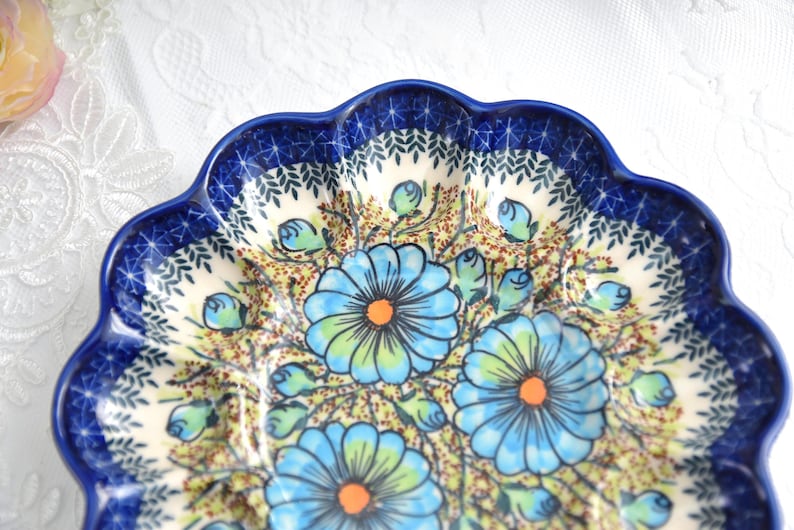 May include: A ceramic plate with a scalloped edge and a floral design. The plate features a dark blue rim with a pattern of leaves and a cream-colored center with blue flowers and green leaves. The flowers have orange centers.