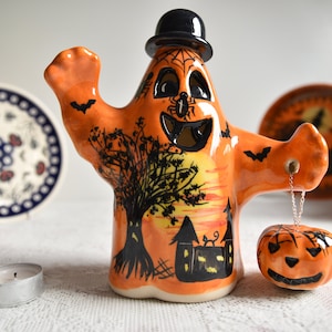 Puede incluir: Una figurita de fantasma de cerámica con un sombrero de copa negro, sosteniendo una pequeña calabaza en una cadena. El fantasma está pintado de naranja con detalles negros, incluyendo una telaraña, murciélagos y un árbol. La calabaza también está pintada de naranja con una cara negra.