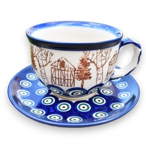 May include: A blue and white ceramic teacup and saucer set with a brown and white design of a house and trees. The saucer has a blue and white eye pattern.