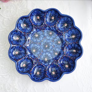 以下が含まれることがあります： 青いセラミック製のサービングプレート、スカラップエッジ付き。 プレートは、青の色合いのフローラルデザインで、赤と白のアクセントが付いています。 中央と外側の縁には、繰り返しの花のパターンがあります。