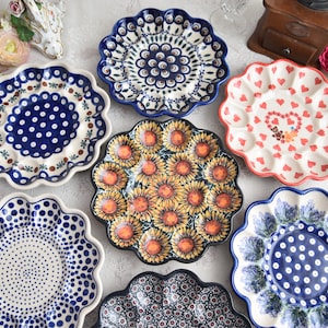 May include: A collection of decorative ceramic plates with scalloped edges. The plates feature various patterns, including sunflowers, hearts, and floral designs in blue, red, and yellow. The plates are arranged on a white surface, showcasing their intricate details and vibrant colors.