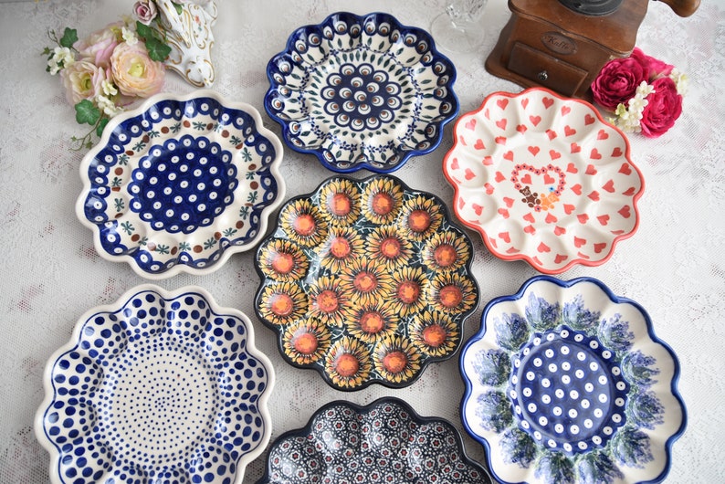 May include: A collection of decorative ceramic plates with scalloped edges. The plates feature various patterns, including sunflowers, hearts, and floral designs in blue, red, and yellow. The plates are arranged on a white surface, showcasing their intricate details and vibrant colors.