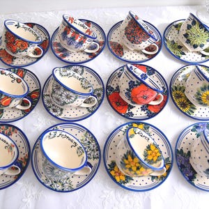 May include: A set of twelve ceramic teacups and saucers, each with a unique floral design and a month's name inside. The cups and saucers have a blue rim and are arranged on a white surface.
