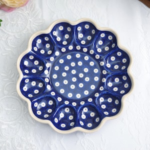 Op de afbeelding: Een blauw en wit keramisch eierserviesbord met een geschulpte rand. Het bord heeft een donkerblauwe achtergrond met witte stippen en een crèmekleurige rand. Het heeft 12 eivormige inkepingen en een centraal cirkelvormig gebied.
