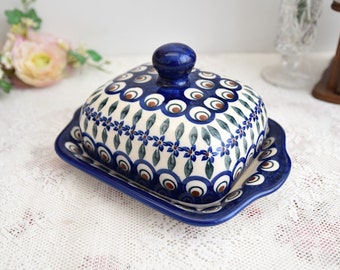 Könnte beinhalten: Eine blaue und weiße Keramik-Butterdose mit Deckel. Die Dose hat ein florales und Pfauenaugen-Design.