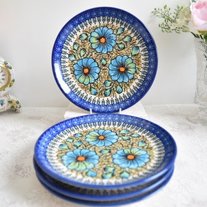 Puede incluir: Cuatro platos de cerámica con un diseño floral azul. Cada plato presenta un borde azul oscuro y un patrón central de flores azules con centros naranjas y hojas verdes. Los platos están apilados sobre una superficie blanca.