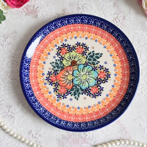 May include: A ceramic plate with a blue rim and a floral design in shades of orange, pink, green, and blue. The center of the plate features a bouquet of three flowers with a white background.