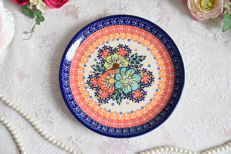 May include: A ceramic plate with a blue rim and a floral design in shades of orange, pink, green, and blue. The center of the plate features a bouquet of three flowers with a white background.