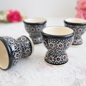 May include: Three black and white ceramic egg cups with a red floral pattern. The cups are on a white lace tablecloth.