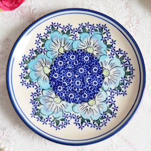 May include: A white ceramic plate with a blue and white floral design. The plate features a central motif of a blue flower surrounded by smaller blue flowers and green leaves. The rim of the plate is decorated with a blue band.
