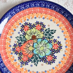 May include: A white ceramic plate with a blue rim and a colorful floral design. The flowers are red, orange, yellow, green, and blue. The plate has a white background with a blue, orange, and yellow pattern around the edge.