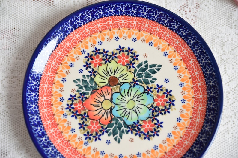 May include: A white ceramic plate with a blue rim and a colorful floral design. The flowers are red, orange, yellow, green, and blue. The plate has a white background with a blue, orange, and yellow pattern around the edge.