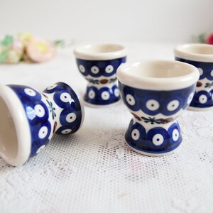 Può includere: Quattro porta uova in ceramica blu e bianca con un motivo floreale. I porta uova sono su un tovaglia di pizzo bianco.