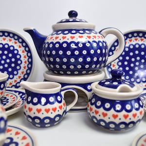 Op de afbeelding: Een blauwe en witte keramische theeset met rode harten. De set bevat een theepot, roomkan, suikerpot en vier theekopjes en schoteltjes.