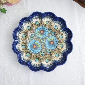 May include: A round, scalloped-edge ceramic plate with a blue floral design. The plate features three large blue flowers with orange centers and green accents, set against a cream-colored background with a dark blue border.
