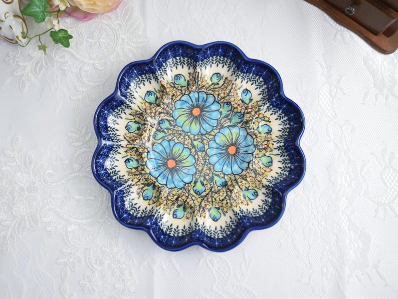 May include: A round, scalloped-edge ceramic plate with a blue floral design. The plate features three large blue flowers with orange centers and green accents, set against a cream-colored background with a dark blue border.
