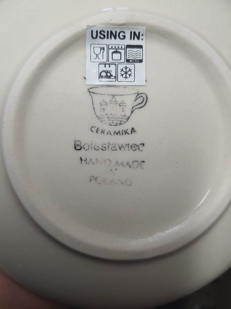 May include: White ceramic saucer with black text that reads "USING IN:" and includes icons for microwave, oven, dishwasher, and freezer safe. The saucer also has black text that reads "CERAMIKA Boleslawiec HAND MADE IN POLAND".