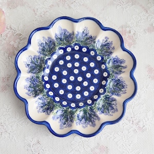 Può includere: Un piatto in ceramica blu e bianco con bordo ondulato. Il piatto presenta un disegno floreale con fiori di lavanda e un motivo centrale a pois.