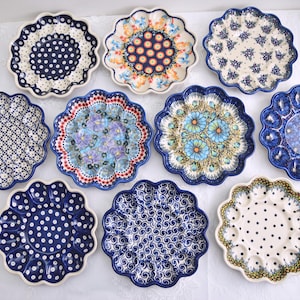 May include: A collection of eleven decorative ceramic plates with scalloped edges. The plates feature various patterns, including polka dots, floral designs, and geometric motifs. The color palette includes blue, white, and touches of other colors.
