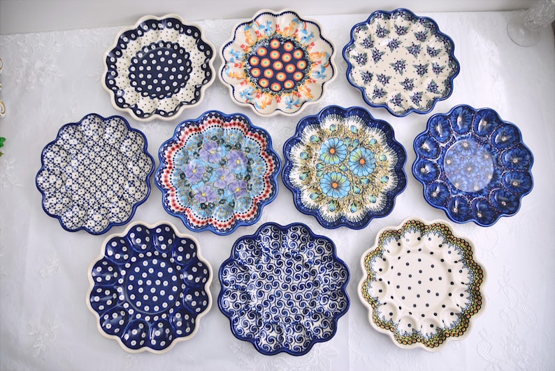 May include: A collection of eleven decorative ceramic plates with scalloped edges. The plates feature various patterns, including polka dots, floral designs, and geometric motifs. The color palette includes blue, white, and touches of other colors.